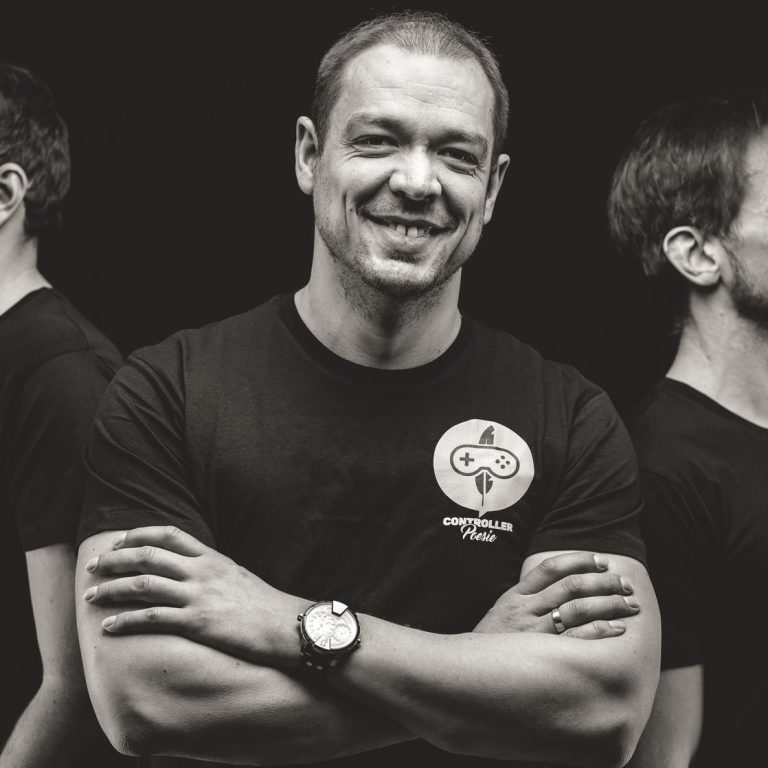 Controller Poesie – Studio-Portrait Spielewelten Foto von Creator Spielewelten mit verschränkten Armen in schwarzem T-Shirt mit Controller-Poesie-Motiv, lächelnd vor dunklem Hintergrund.